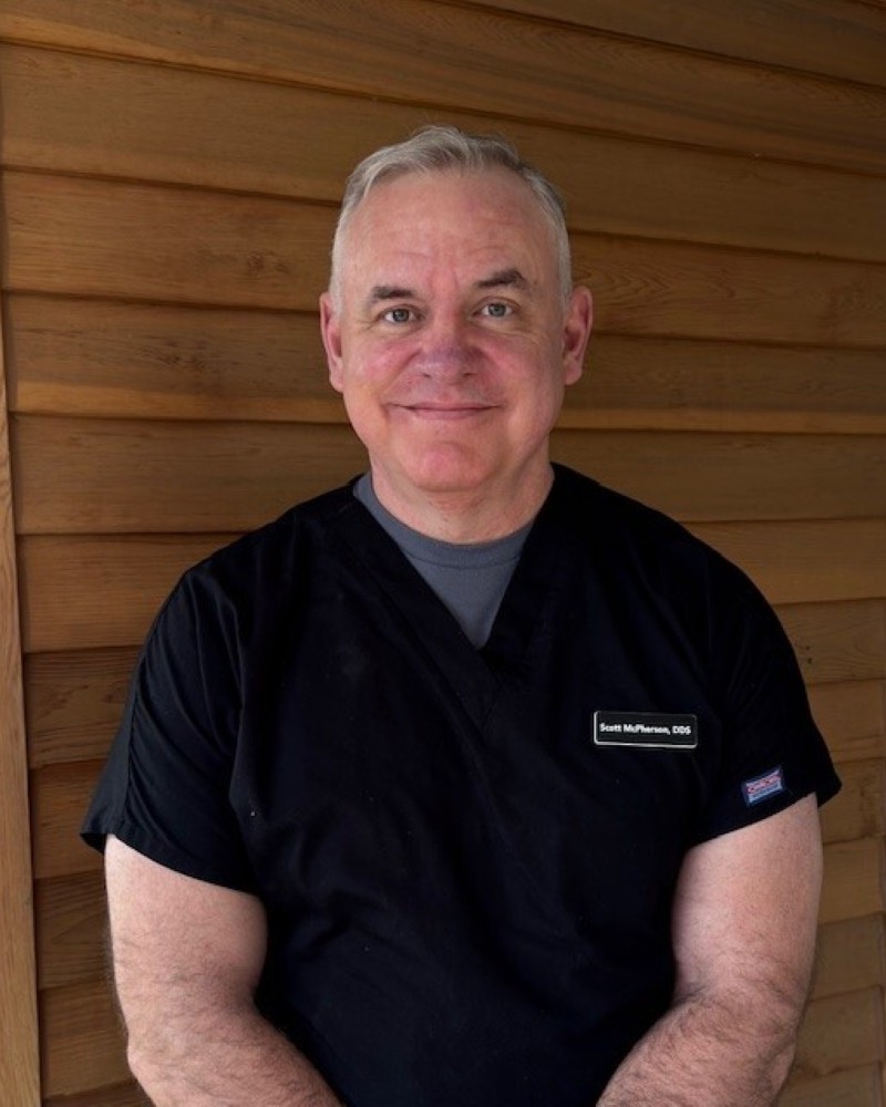 Dr. Scott McPherson, DDS, smiling in dental scrubs against a wooden background, representing Hickman Family Dental's experienced dental team.